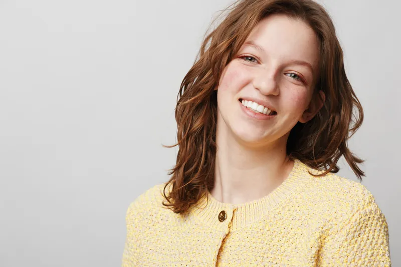 Smiling young woman with brown hair wearing yellow knitted sweater looks at camera on grey background. Portrait of happy female model with natural makeup and casual style in studio lighting.