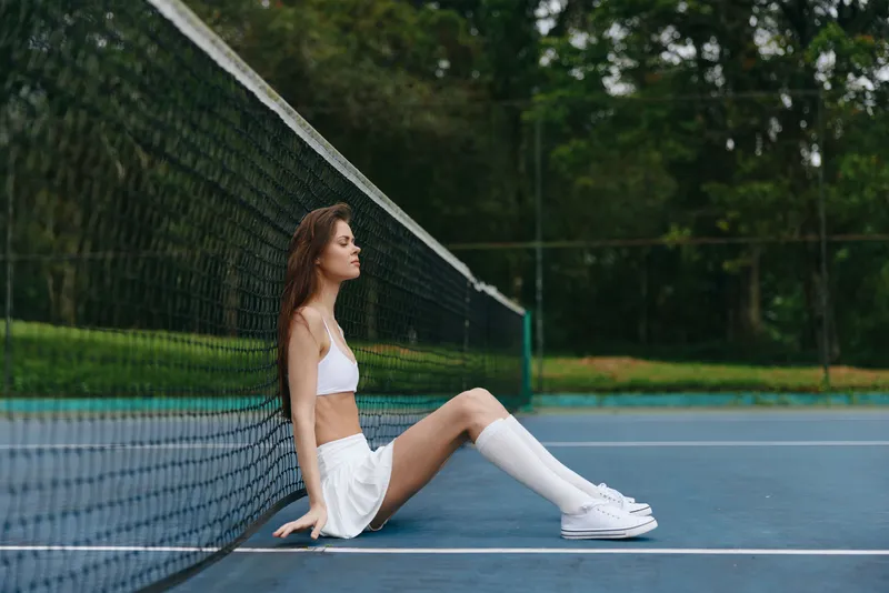 Young athletic woman in white sportswear relaxing on the tennis court, exuding confidence and determination in a lush green environment