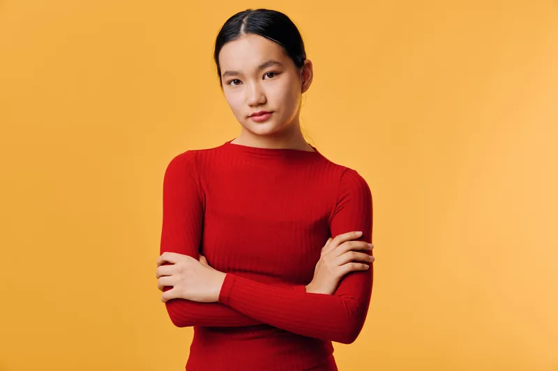 Young woman in red sweater stands with crossed arms against orange background, looking confidently at camera with calm expression and straight posture in studio portrait.