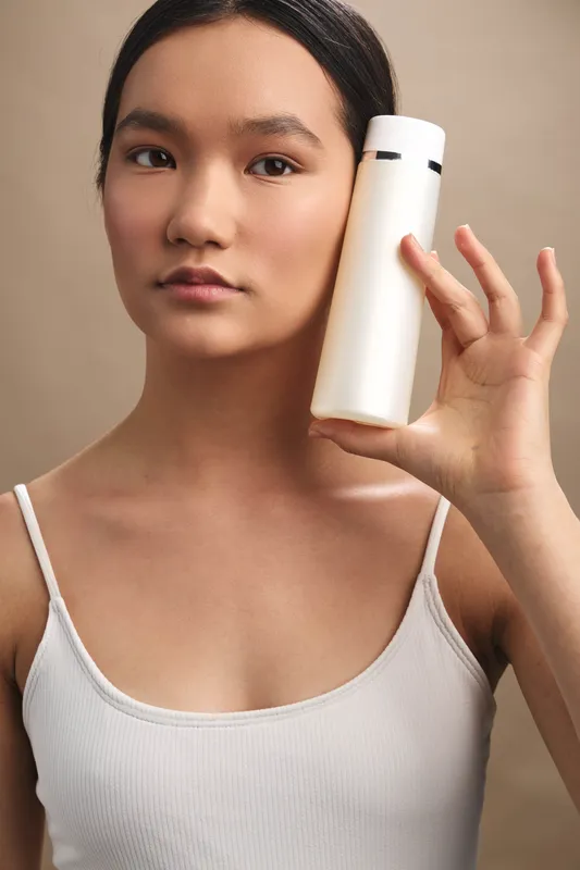 asian woman holds a white bottle in a simple studio setting, focused expression, beauty product shot. clean background and soft lighting convey minimal elegance, modern cosmetic presentation,