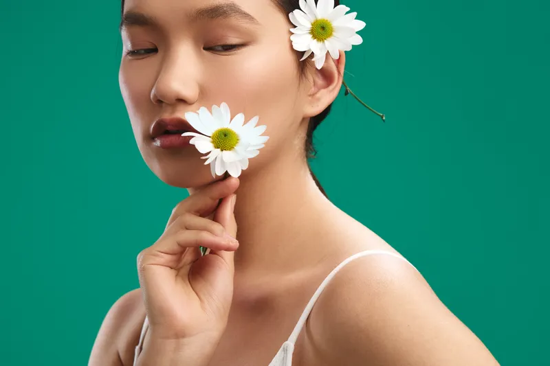 Asian woman with flowers in her hair stands against a green backdrop, soft light highlights smooth skin and a serene expression, creating a fresh, elegant beauty in a natural portrait.