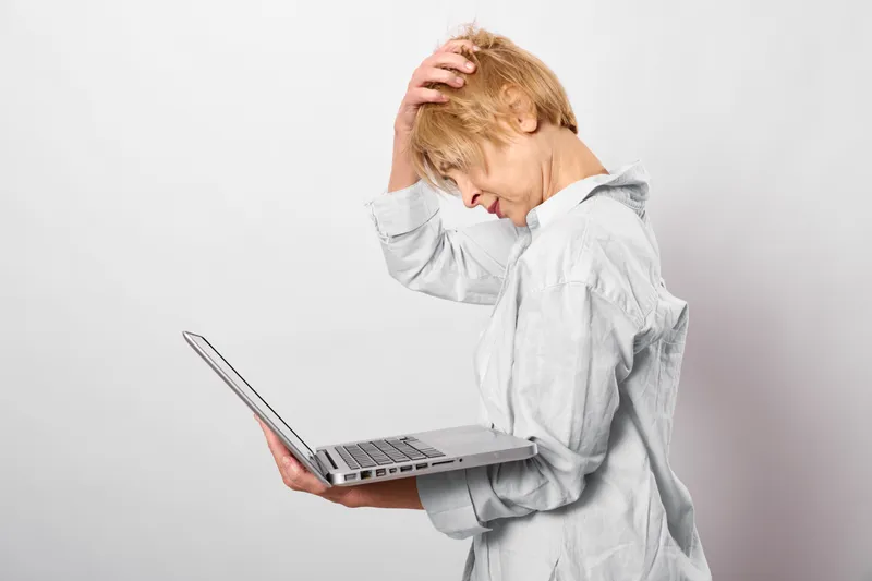 Overwhelmed elderly woman shows anxiety and stress, holding her head beside a laptop. Concept of burnout and mental health.