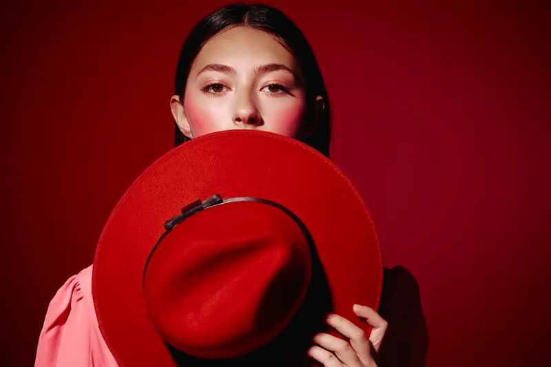 Fashionable young woman with dark hair holds large red hat in front of her face. Studio portrait on red background. Concept of beauty, style, fashion, and complement creates mysterious mood.