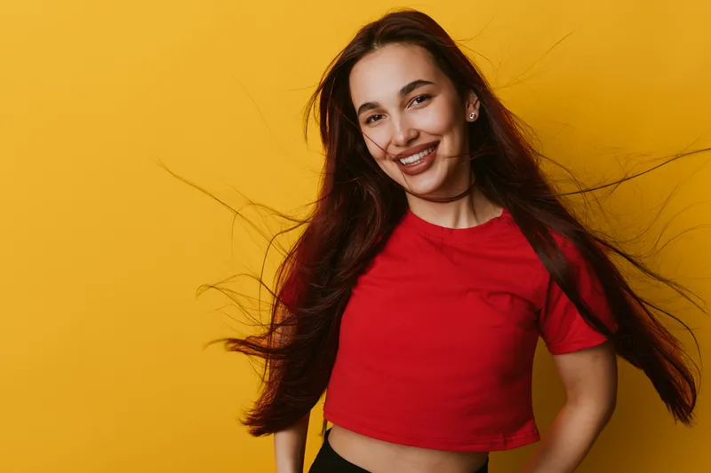 Young woman with long hair smiling and wearing red shirt standing against yellow background. Portrait of happy female with natural look and casual style in studio setting.