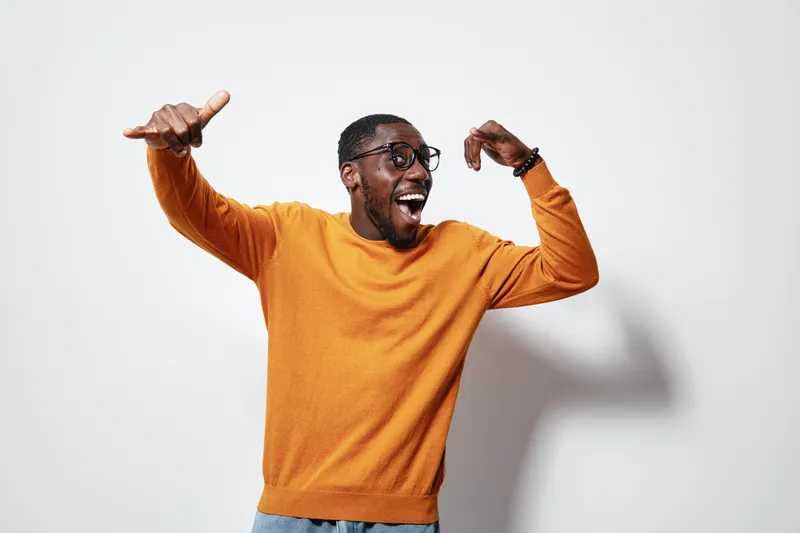 African American man with dark skin wearing orange sweater and glasses posing excited with thumbs up and smile on white background. Casual style and joyful expression.
