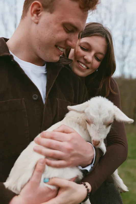 Loving couple outdoors gently holds a friendly goat kid, sharing warm smiles in a rural setting, capturing everyday lifestyle and connection with animals, friends nearby in soft natural light