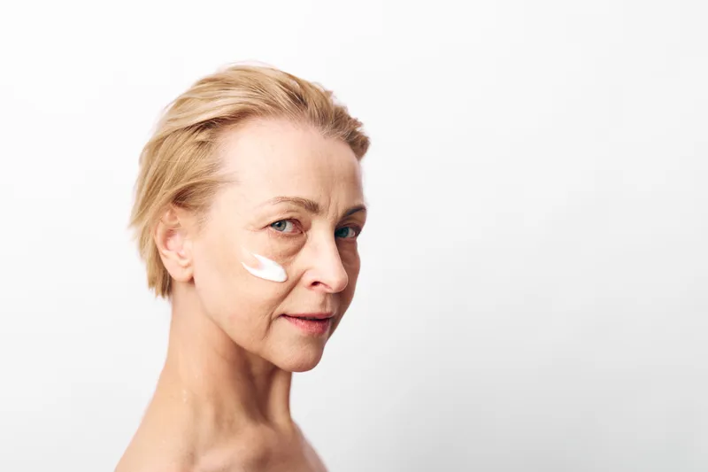 Mature woman with short blonde hair applies white face cream on cheek against white background. Concept of skincare, antiaging, selfcare, natural beauty, and facial wellness.