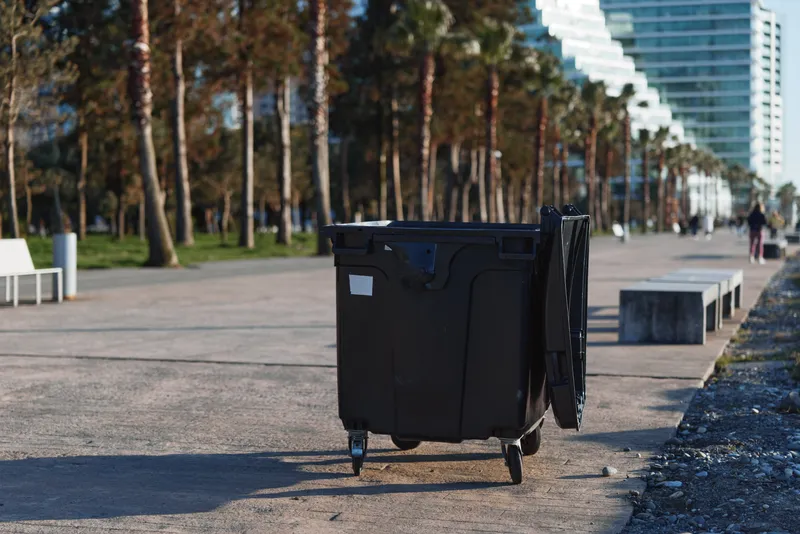 Lone black trash can on a paved promenade, trees and modern buildings in the background, daytime urban scene with a calm, functional atmosphere