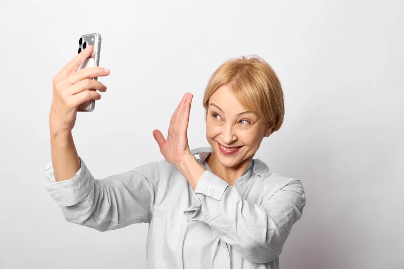 Elderly woman takes a selfie with smartphone, smiling warmly on a white studio background, playful peace gesture, portrait framing, concept of joyful aging and self presentation