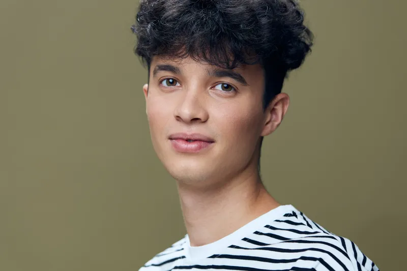 young man with curly hair wearing striped shirt, looking at camera with calm expression against olive background, natural light portrait in casual style