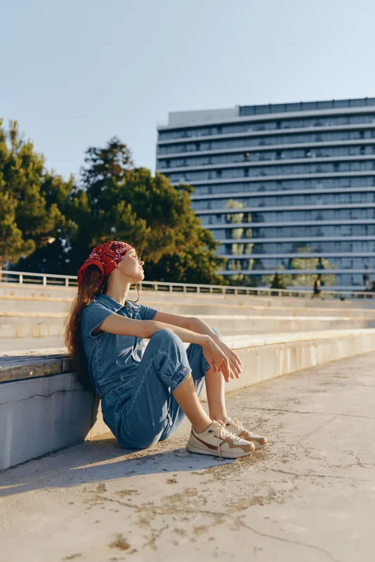young woman sitting outdoors on concrete steps wearing denim outfit and red bandana during sunny day with urban background