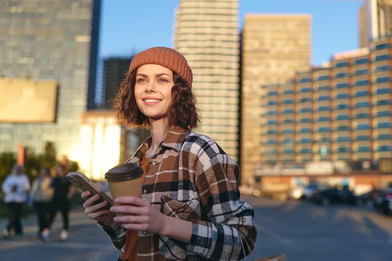 Woman holding coffee and phone in a city street at golden hour, smile bright and candid, authenticity and mindful living captured in warm golden glow, urban lifestyle and emotional storytelling.