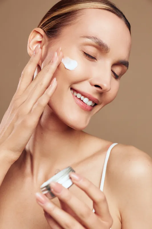 Young woman applies moisturizer to her cheek, smiling softly as she dispenses cream from a jar, isolated against a light beige backdrop.
