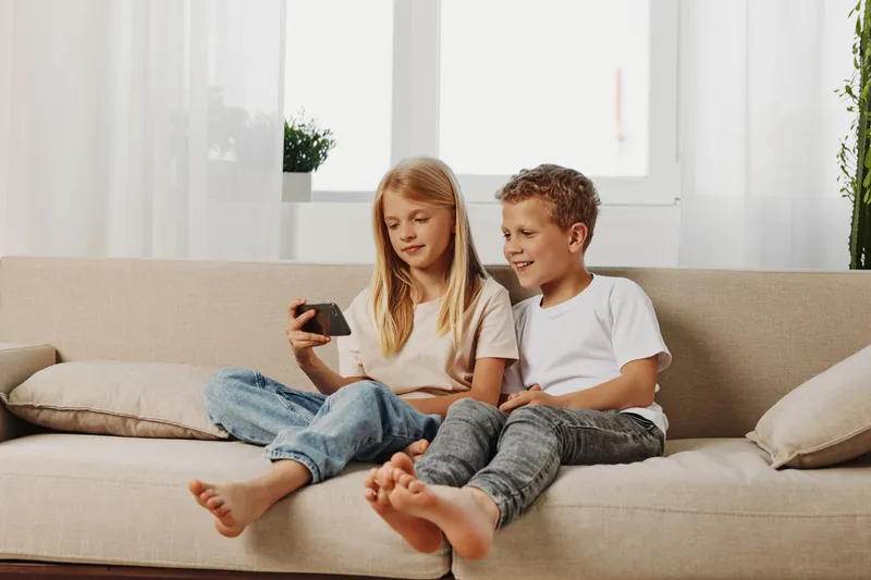 Two children sitting barefoot on a beige sofa looking at a smartphone together in a bright living room. Casual home setting with natural light and relaxed atmosphere.
