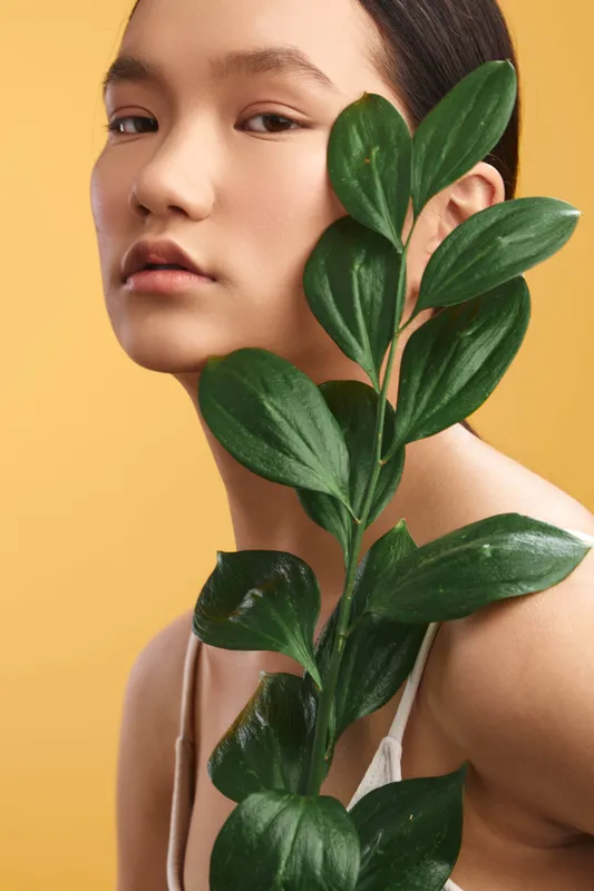 Asian woman gazes softly at the camera with a leafy plant beside her, creating a fresh, natural mood and serene beauty in a studio portrait.