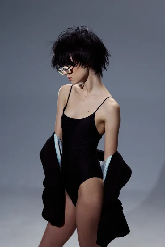 Slim woman in black bodysuit and glasses posing with black jacket in studio with gray background.