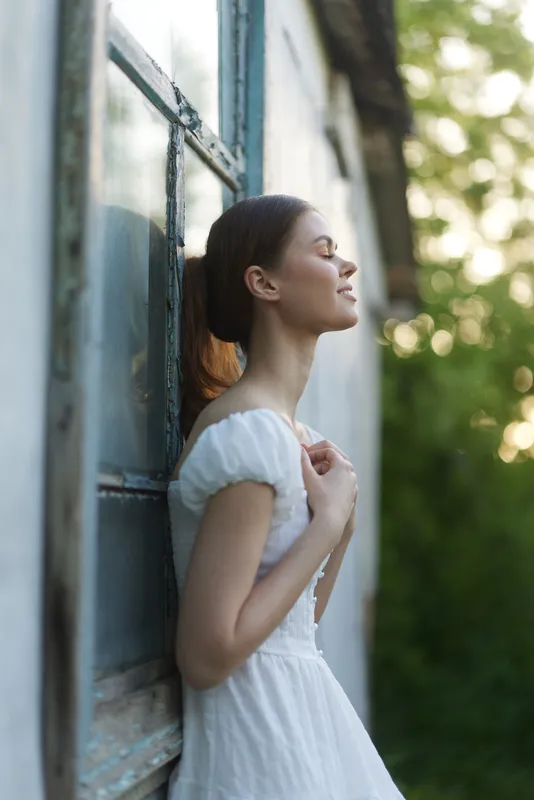 Young woman embracing nature, wearing a white dress, with a peaceful expression by the window, surrounded by greenery and soft natural light