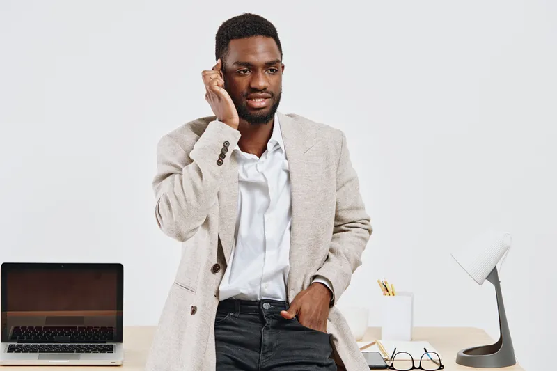 Professional-looking young man in stylish beige blazer and white shirt standing at modern office desk, smiling confidently, talking on mobile phone, with laptop and office accessories on background. B