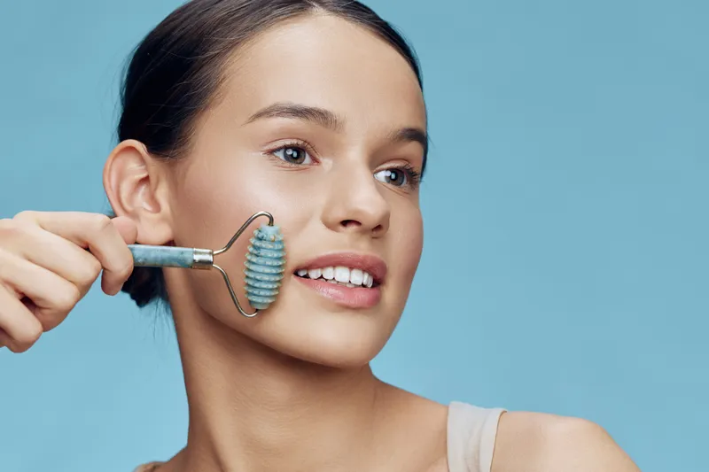 Young woman using a jade roller for facial massage on a light blue background, showcasing healthy skin and relaxing beauty routine, emphasizing wellness and skincare practices