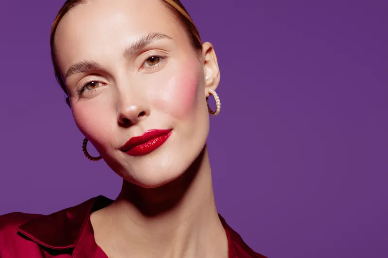 Close-up portrait of a confident woman with red lipstick and hoop earrings against purple background. Concept of beauty, fashion, makeup, and elegance in studio lighting.