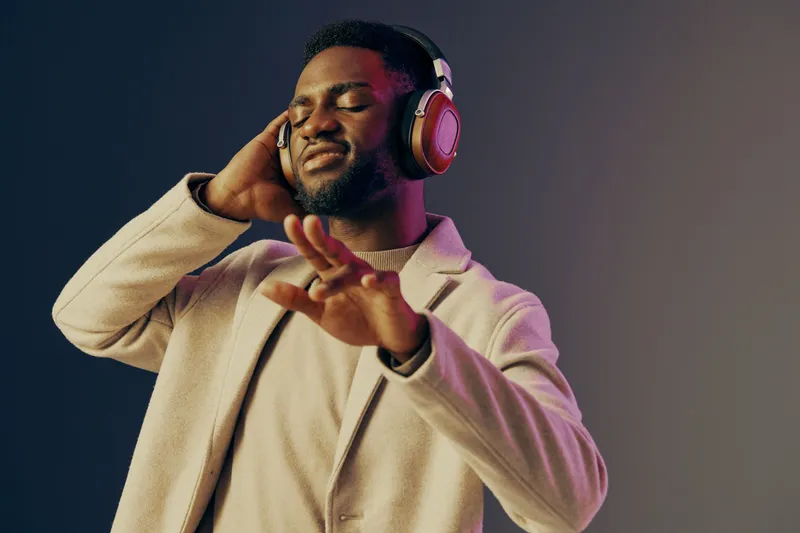 A joyful young Black man enjoying music with headphones, wearing a stylish cream jacket, set against a gradient background, conveying relaxation and happiness