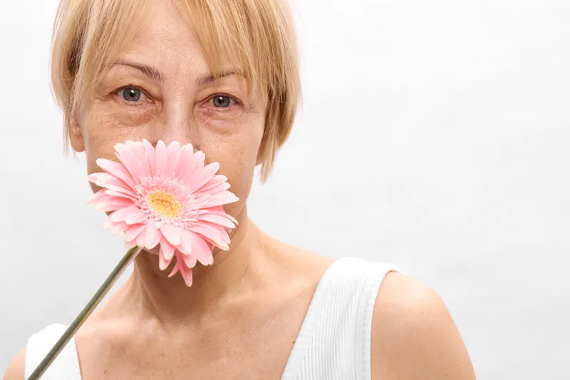 Blonde woman portrait with pink gerbera flower covering lips, soft white background, gentle beauty and spring mood, concept of selfcare and calm femininity today