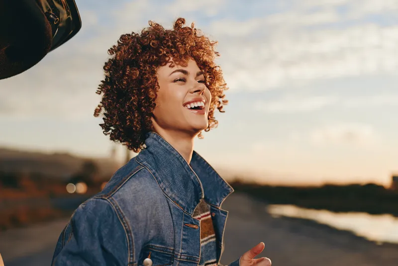 Smiling woman with curly hair wearing a denim jacket outdoors at sunset, warm golden light, relaxed peaceful lifestyle mood, scenic hills and road behind, soft breeze and open sky