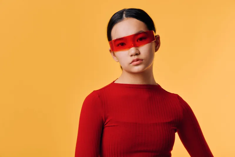 Young woman with red sunglasses and red sweater posing in studio with orange background. Fashion portrait of confident model with sleek hairstyle and modern eyeglasses accessory.