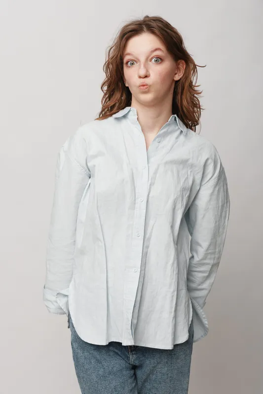 Young woman with curly hair wearing white shirt and jeans makes funny face with pursed lips and raised eyebrows. Casual style, playful mood, isolated on light background.