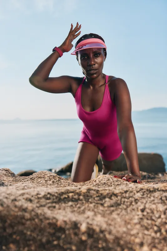 На изображении запечатлена уверенная женщина в розовом купальнике, стоящая на коленях на каменистой местности у моря