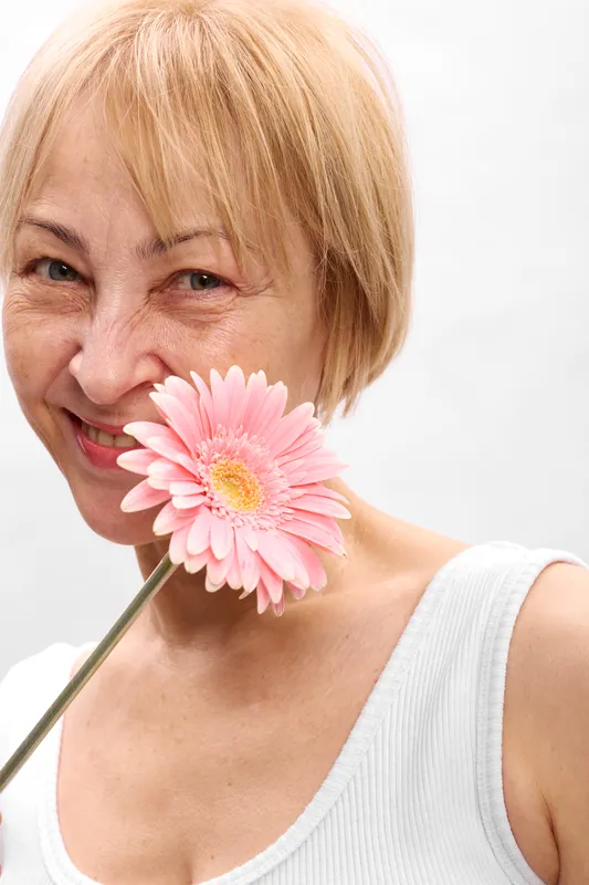 Portrait of a smiling senior blonde woman on a clean white studio background, holding a pink gerbera flower near her lips in a close up headshot soft.