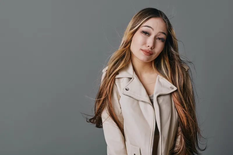 Young woman with long hair and natural makeup wearing beige jacket posing with gentle smile on gray background. Beauty, fashion, casual style and confident look closeup portrait.