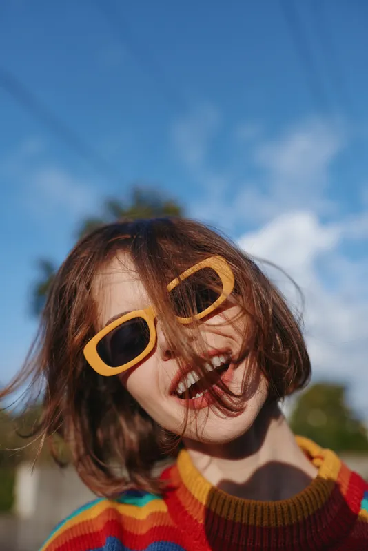 Woman smiling in yellow sunglasses and striped sweater, short hair portrait outdoors under blue sky, playful fashion lifestyle shot with joyful travel vibe and sunlit face.