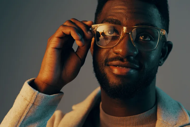 Young Black man adjusting his glasses, smiling confidently in a stylish outfit against a modern gradient background KEY glasses, young, man, smiling, stylish, modern, confidence, fashion, gradient,