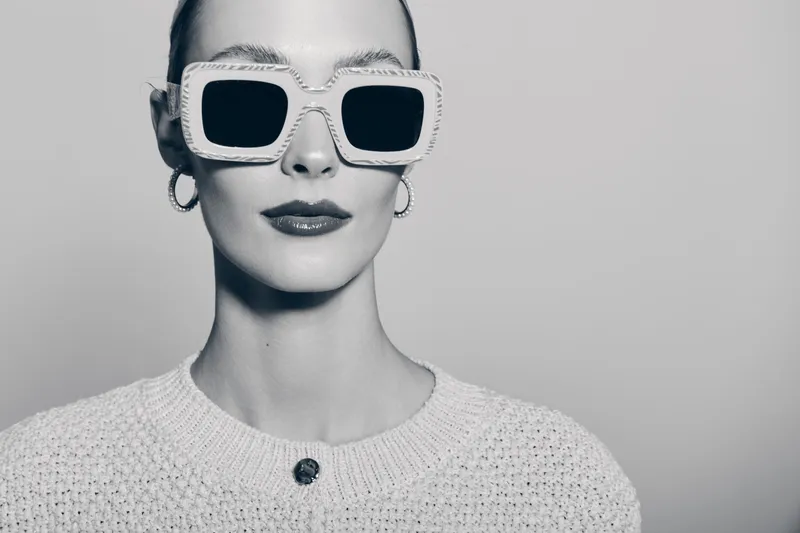 Stylish white woman with fair skin wears large square sunglasses and knitted sweater, posing confidently against monochrome background. Concept of fashion, accessories, and modern elegance.