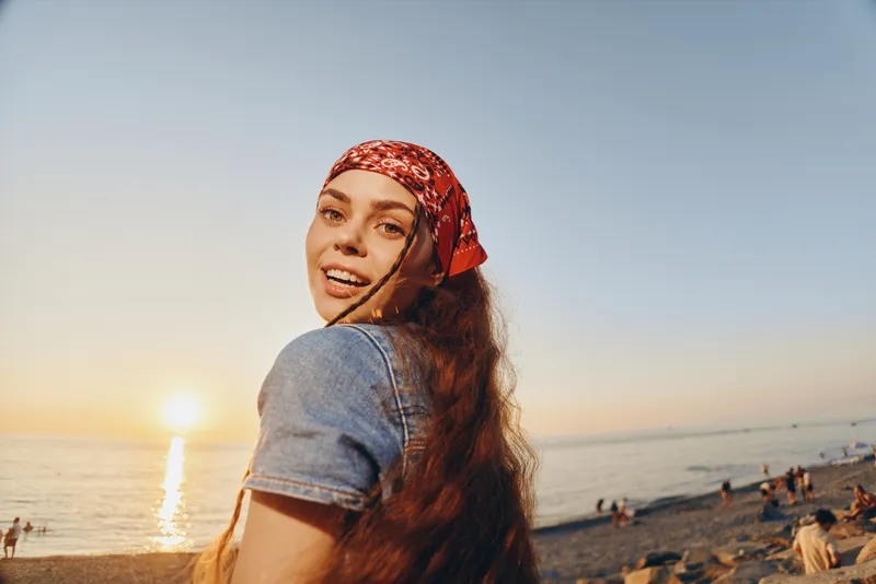 smiling woman wearing red headscarf and denim jacket enjoying sunset on beach with calm sea and clear sky background