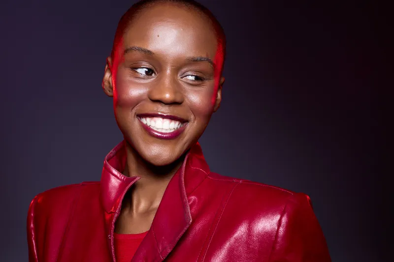 Smiling African woman with dark skin wearing a shiny red leather jacket looks to the side against a purple background. Concept of fashion, confidence, and beauty with vibrant colors.
