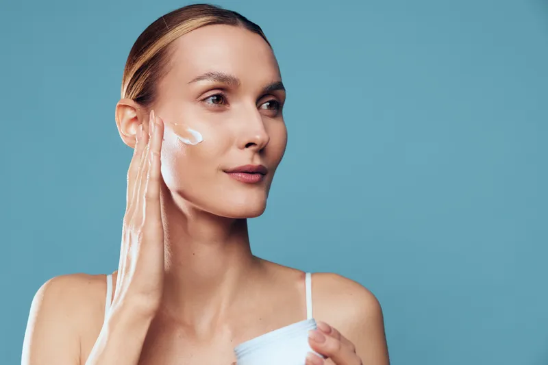 Young woman skincare routine, applying facial cream with moisturizer on cheeks, holding small bottle, calm beauty concept, smooth skin, studio shot, blue background, isolated clean look