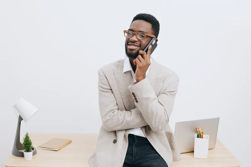 Friendly smiling man with glasses talking on mobile phone while standing at modern office desk, dressed in smart casual attire, with laptop and office supplies. Work and communication concept.