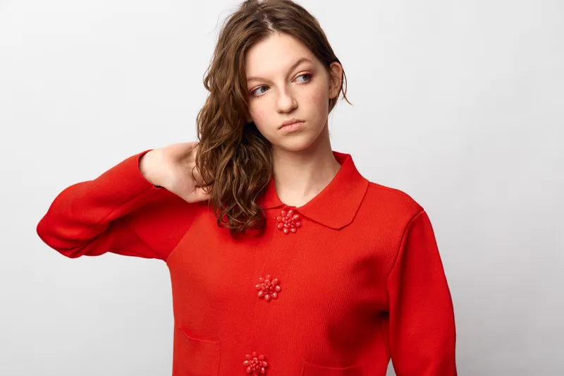 Young woman in red sweater looks away with thoughtful expression, hand behind head. Studio portrait on white background. Concept of contemplation, uncertainty, and casual fashion.