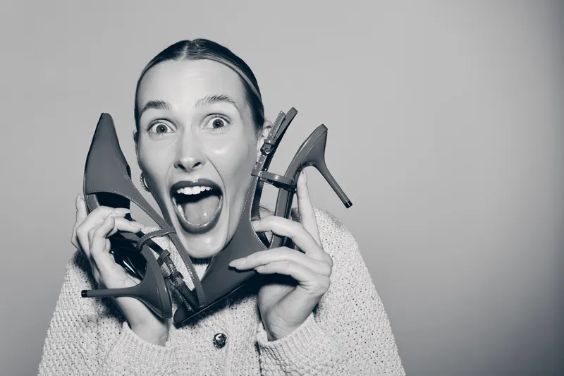 Happy Caucasian woman with fair skin holds high heel shoes near face smiling widely. Concept for fashion, footwear, joyful emotions and shopping.