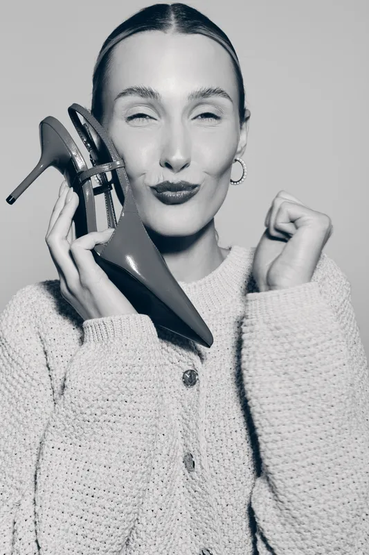Young white woman with fair skin holds high heel shoe near face, smiling with closed eyes. Casual knitted sweater worn, studio portrait in black and white. Concept for fashion and lifestyle.