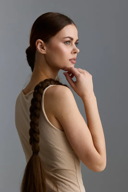 thoughtful woman with long braided hair and sleeveless beige top posing against neutral gray background, showcasing natural beauty and calm expression