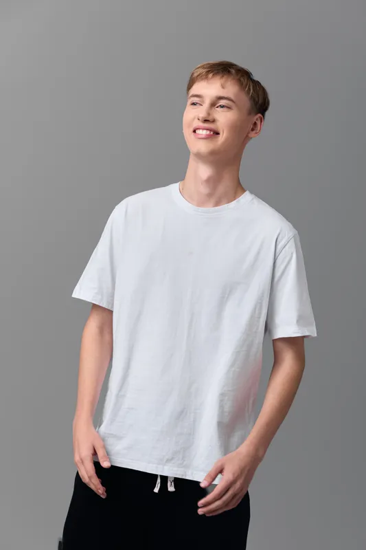 Young man wearing a white t shirt poses with relaxed style in a studio, gray backdrop, casual wear vibe, smiling lightly, showcasing comfortable everyday fashion and confident mood
