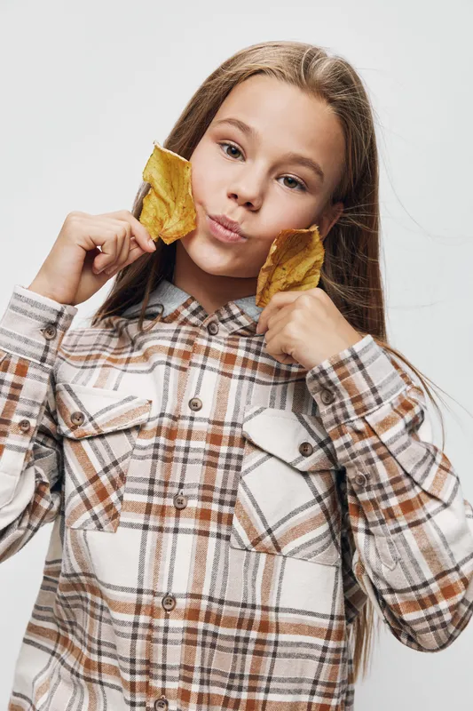 Girl with long hair in plaid shirt holding two yellow autumn leaves near face with playful expression on light background. Seasonal, casual, youth and nature concept.