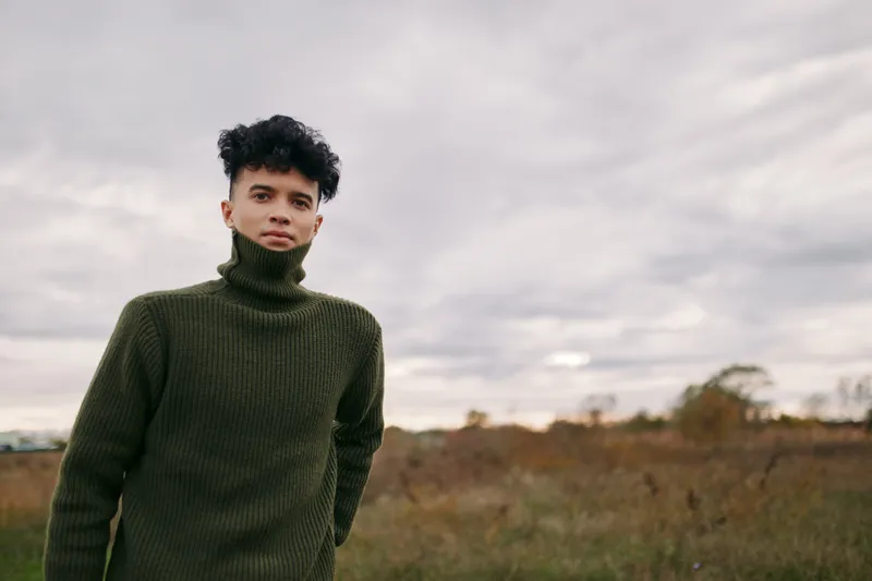 Authenticity and veracity guide a candid portrait of a young man in a dark green knit turtleneck, standing in a bare field under overcast skies, with honest detail and quiet presence.