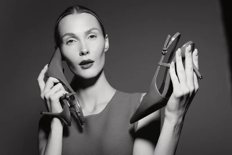 Slim white European woman with fair skin holds red high heels near face in studio. Fashion concept with shoes and model in black and white photography.
