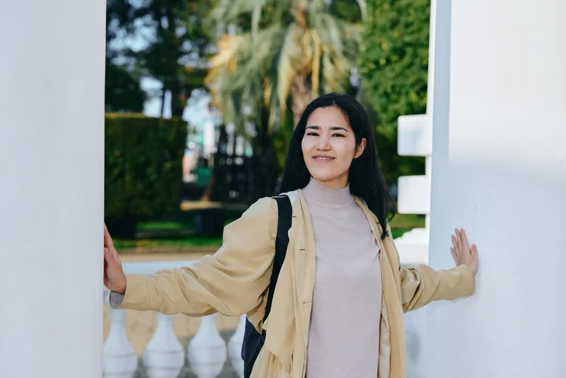 Smiling woman stands between white arches outdoors, wearing a light jacket and backpack, relaxed pose under bright daylight in a park, conveying casual style, warm mood, and welcoming, everyday