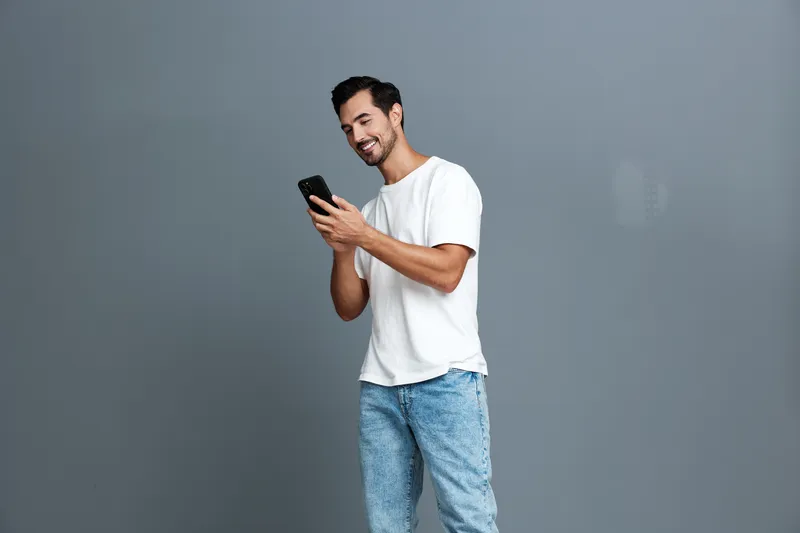 Smiling young man wearing a casual white t-shirt standing while using his mobile phone, exuding a joyful and relaxed vibe against a neutral gray background.