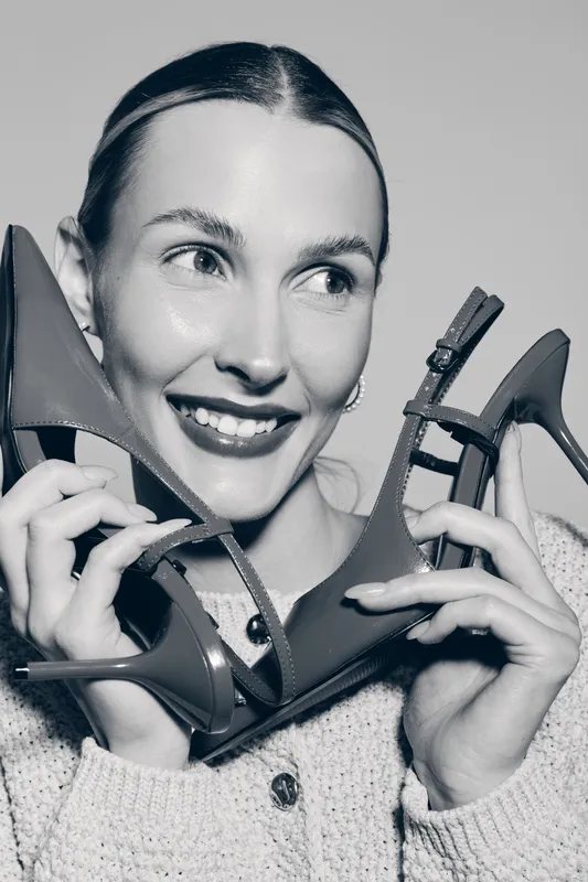 Smiling Caucasian woman with fair skin holds high heel shoes near face, joyful expression, studio portrait. Concept of fashion, happiness, and footwear.